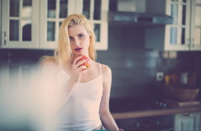 Sensual beauty blonde girl Takha eating an apple with white top in the kitchen